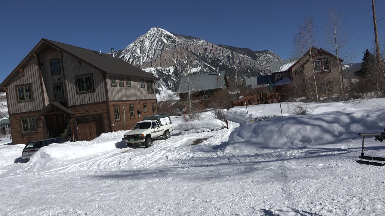 Second frame shows the Crested Butte street with packed snow in front, the white car slightly left of center, houses along the road, a snowy mountain in back, and no visible people.