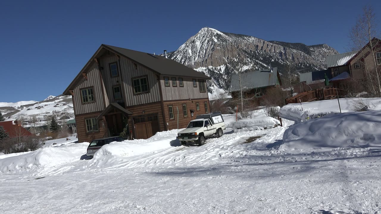 A similar early view in Crested Butte, Colorado shows the white truck centered below the tan and brown house, with packed snow tracks and the mountain ridge behind, and no visible people.