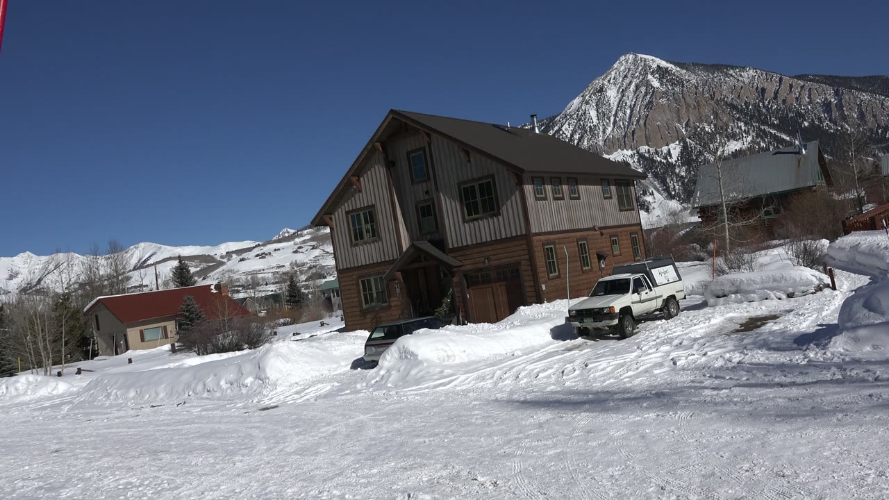 Snowy brown house with a white truck and mountain ridge behind it.