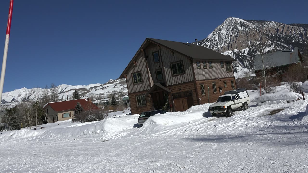 Crested Butte frame with the brown house centered, the parked white truck at right, packed snow in front, distant houses at left, and no visible people.