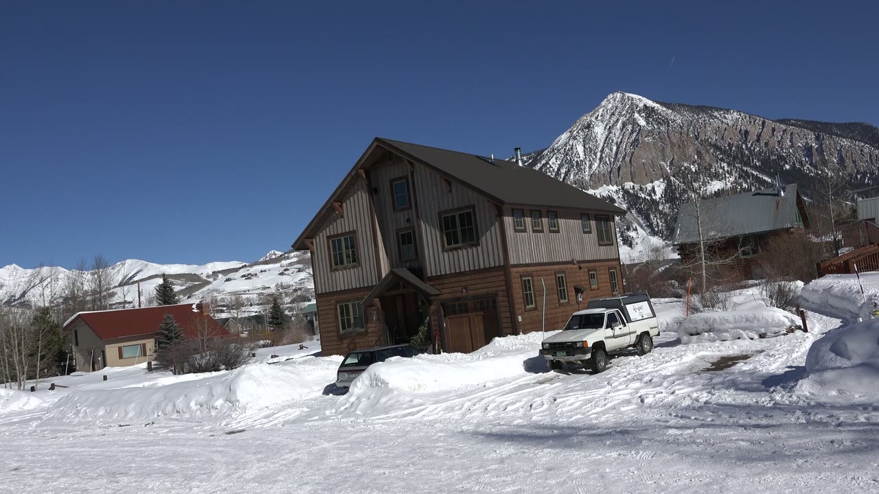 Snowy cabin, parked white vehicle, and mountain backdrop under blue sky.
