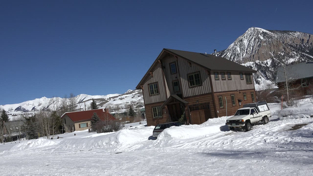 A tilted ground-level view in Crested Butte, Colorado shows a tan and brown building beside plowed snow, a white SUV, and snow-covered mountains, with no visible people.
