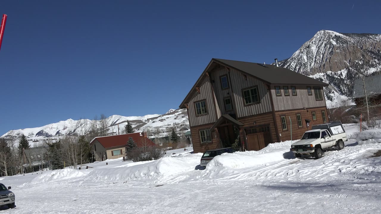 A Crested Butte winter frame shows the tan building and white SUV shifted slightly right, with a tall red and white roadside pole rising near the snowy mountain backdrop and no visible people.