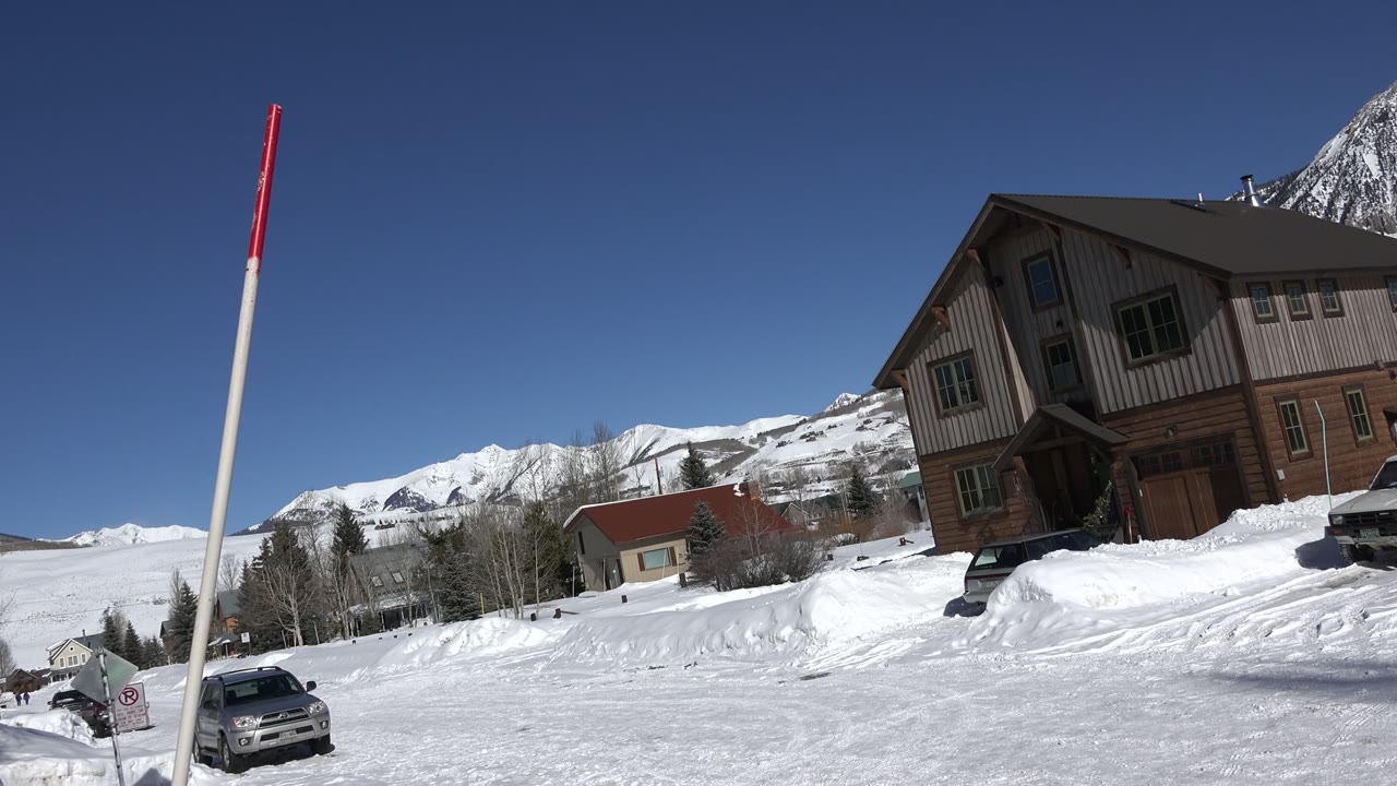 Snowy road with marker pole, parked car, houses, and mountains.