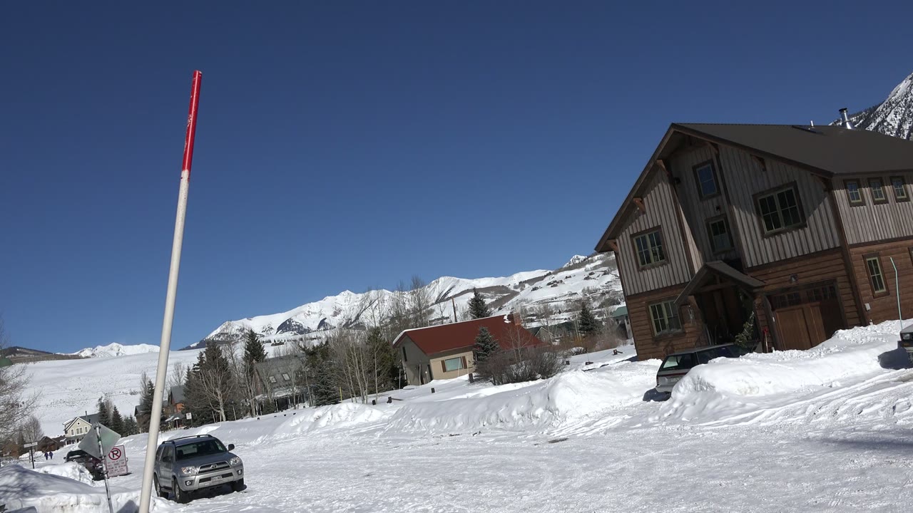 At 0:01 in Crested Butte, Colorado, the view holds on a tilted snowy street with the marker pole near center, a parked car below it, houses to the right, mountains behind, and no visible people.