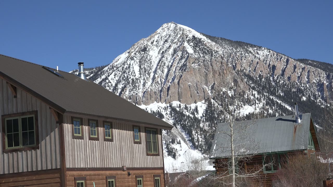 Crested Butte, Colorado frame with the mountain peak centered above a dark roof, a metal-roofed building, bare trees, and no visible people.