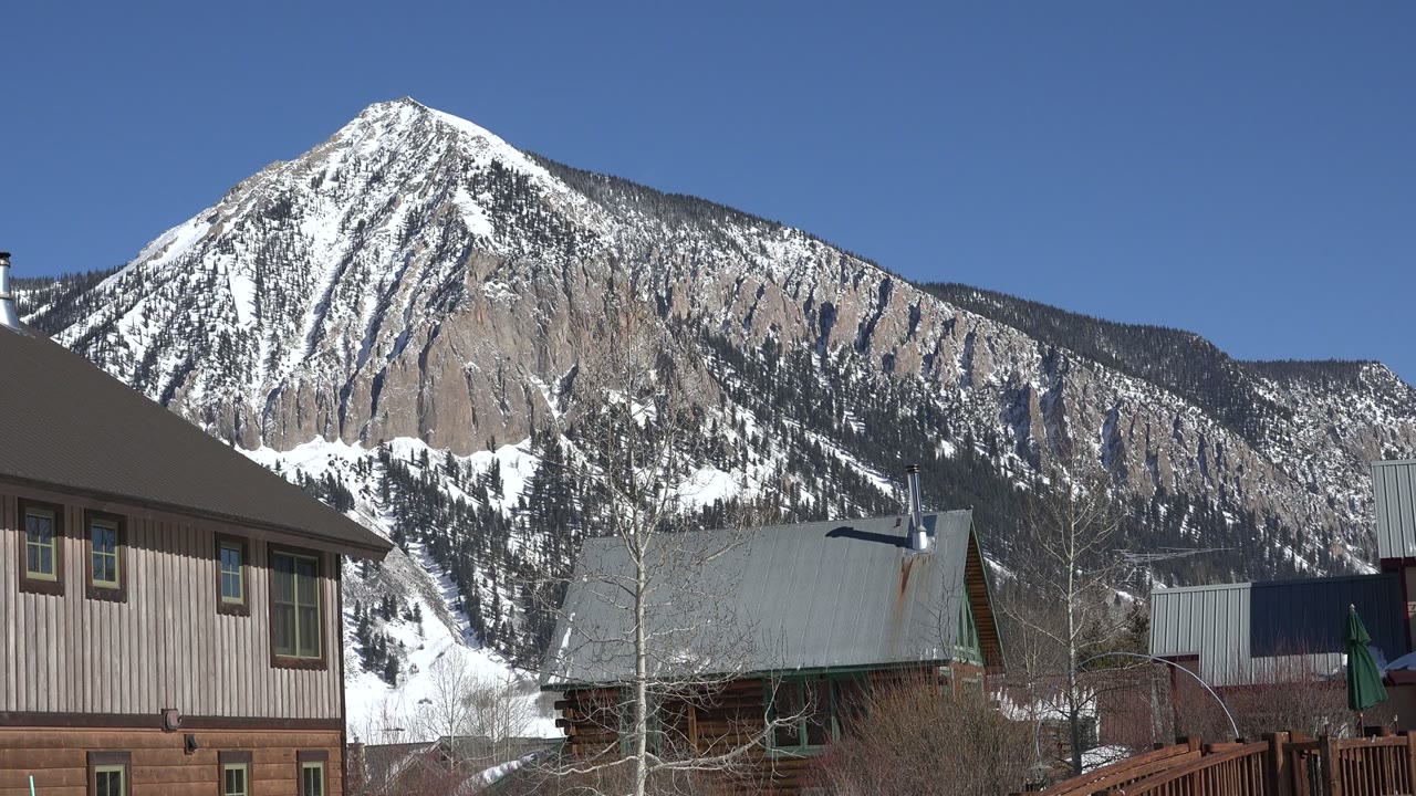 The next frame keeps the snow-striped mountain centered above wood buildings, a greenish metal roof, bare trees, and blue sky in Crested Butte, Colorado, with no visible people.