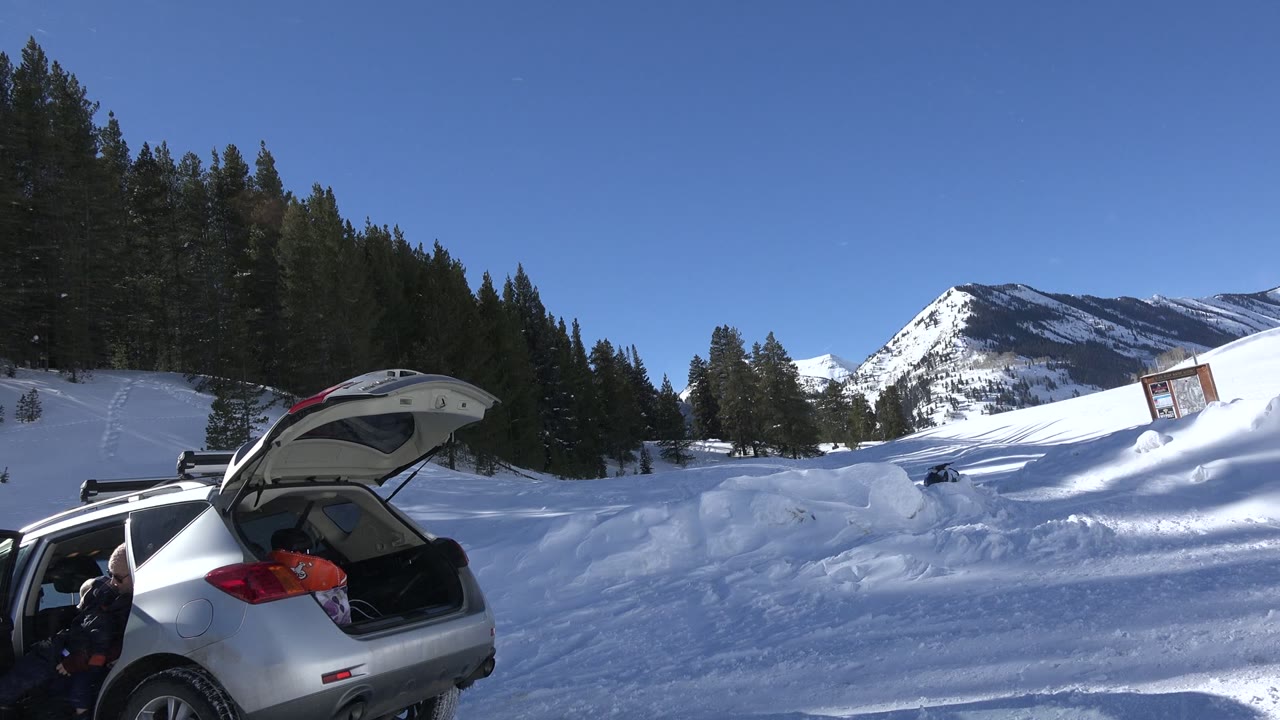 Silver hatchback with its rear liftgate open at the edge of a snowy Crested Butte mountain scene, with evergreen trees, blue sky, and a partly visible person near the car.