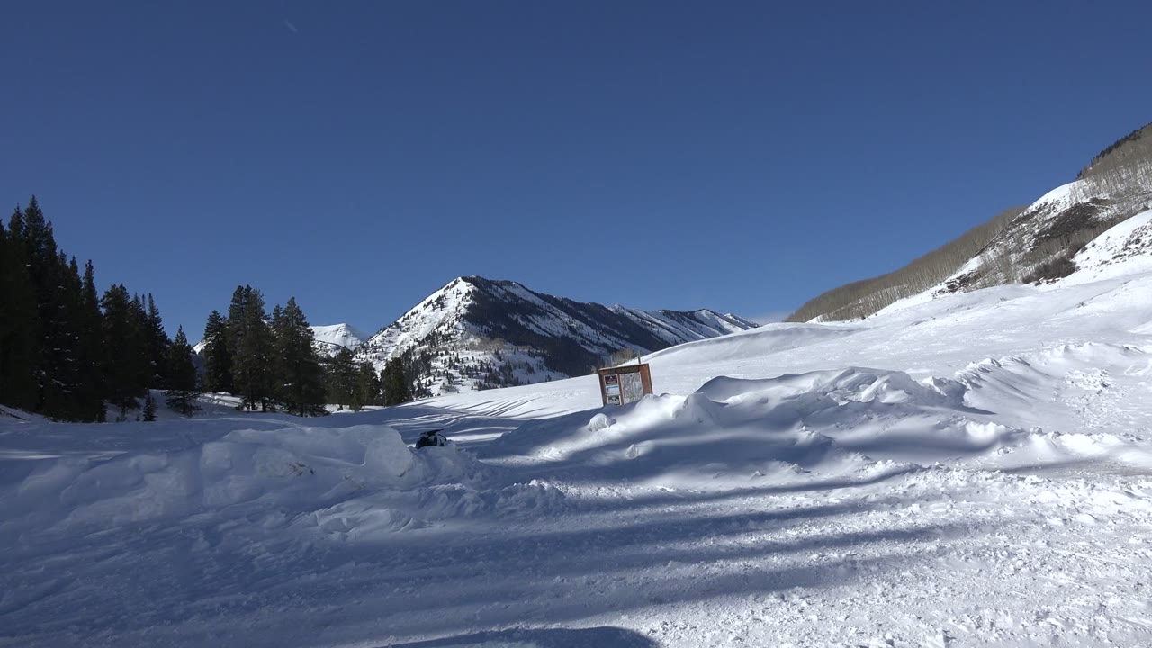 Opening view of snow-covered mountains in Crested Butte, Colorado, with packed snow in front, pines at left, a small sign near center, and no visible people.