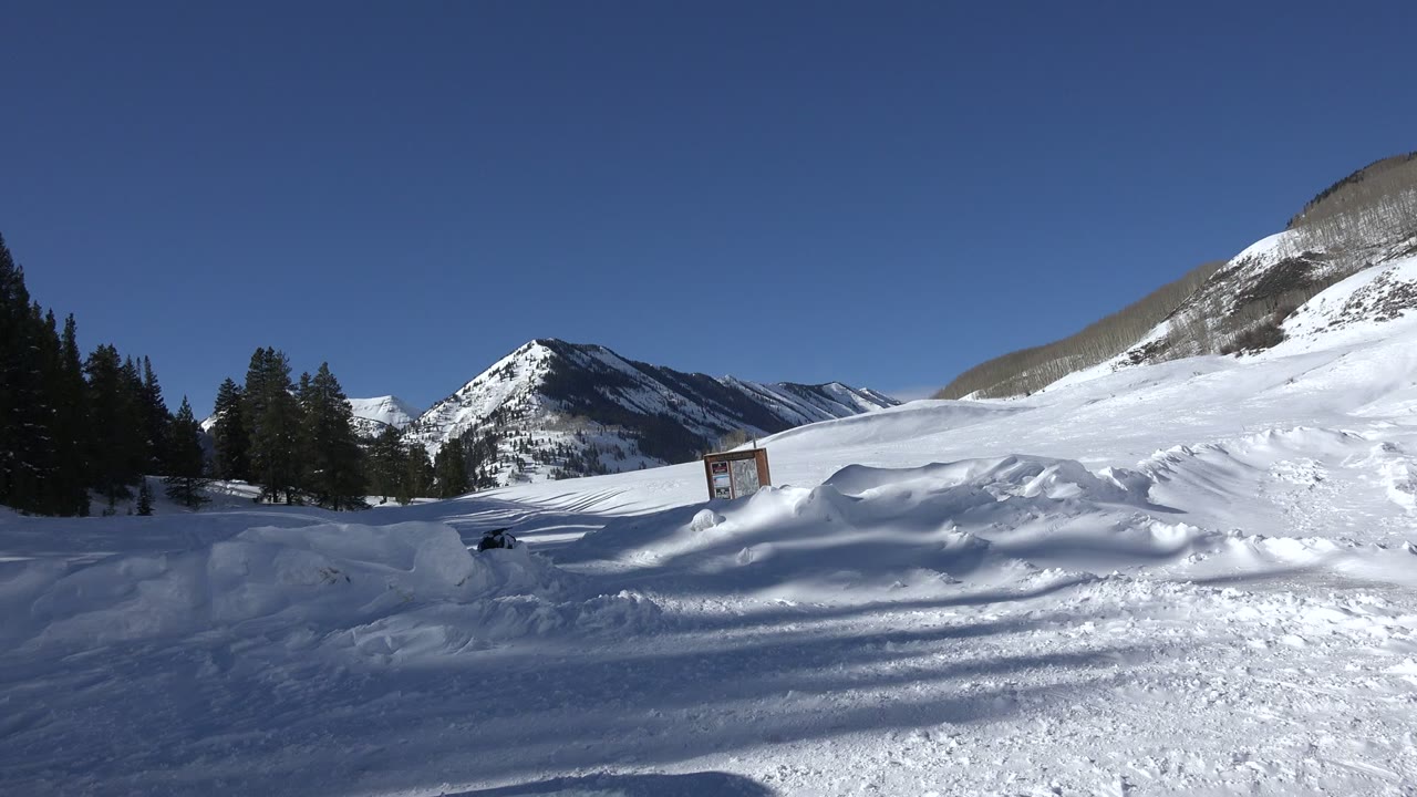 Early wide frame of the Crested Butte winter landscape, showing rippled snowbanks, dark pines, distant ridges, a small posted sign, and no visible people.