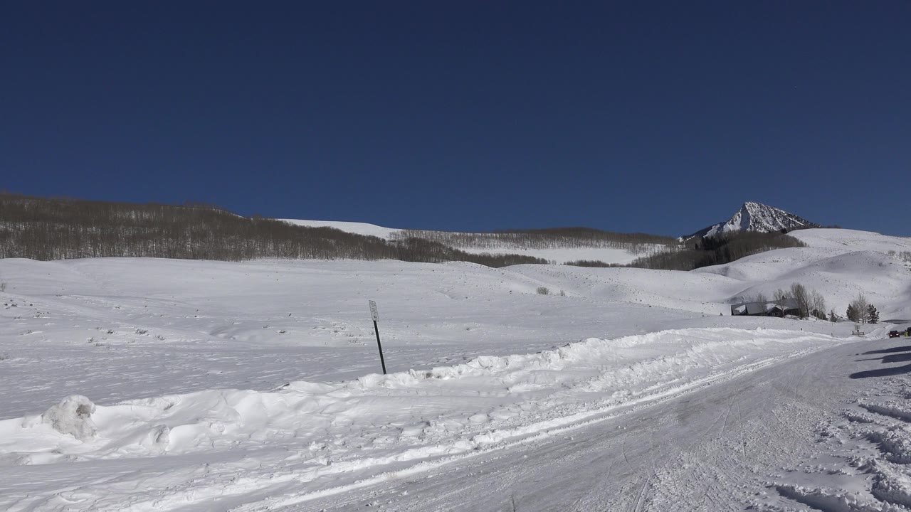 A Crested Butte, Colorado winter road covered in snow and ice runs past open white fields toward a mountain ridge, with no visible people.