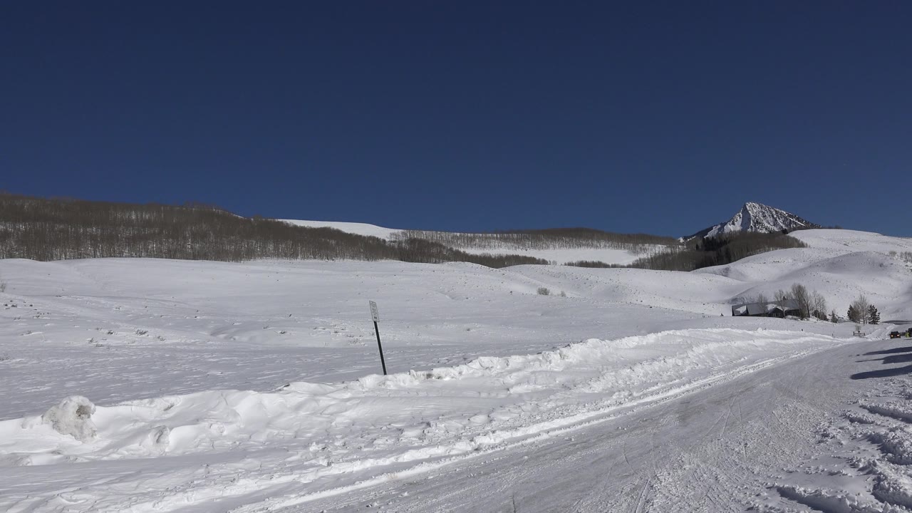 The road in Crested Butte curves through plowed snowbanks with a single roadside post, distant hills, a pointed mountain peak, and no visible people.