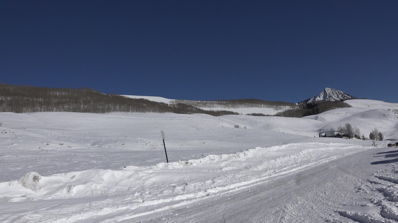 Wide snowy road scene with fields, hills, and a mountain peak under blue sky.