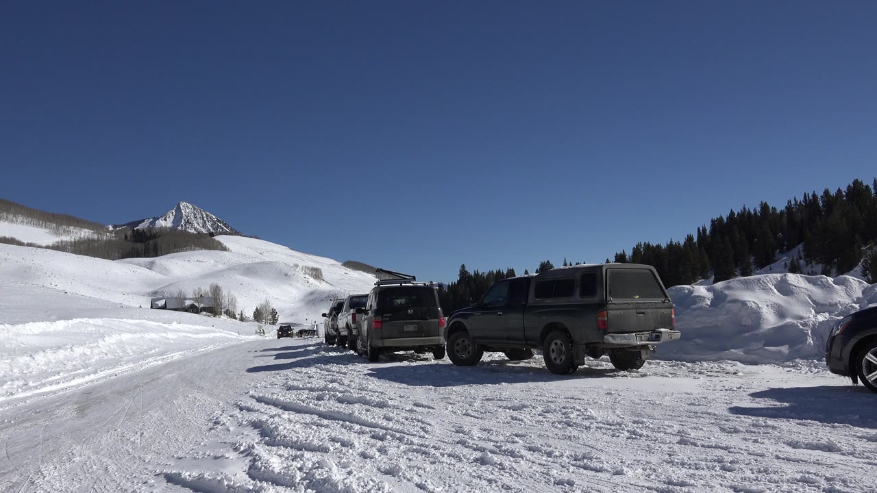 Opening frame of a snow-covered road in Crested Butte, Colorado with parked trucks at right, plowed snowbanks, white hills, evergreen trees, a pointed mountain, and no visible people.