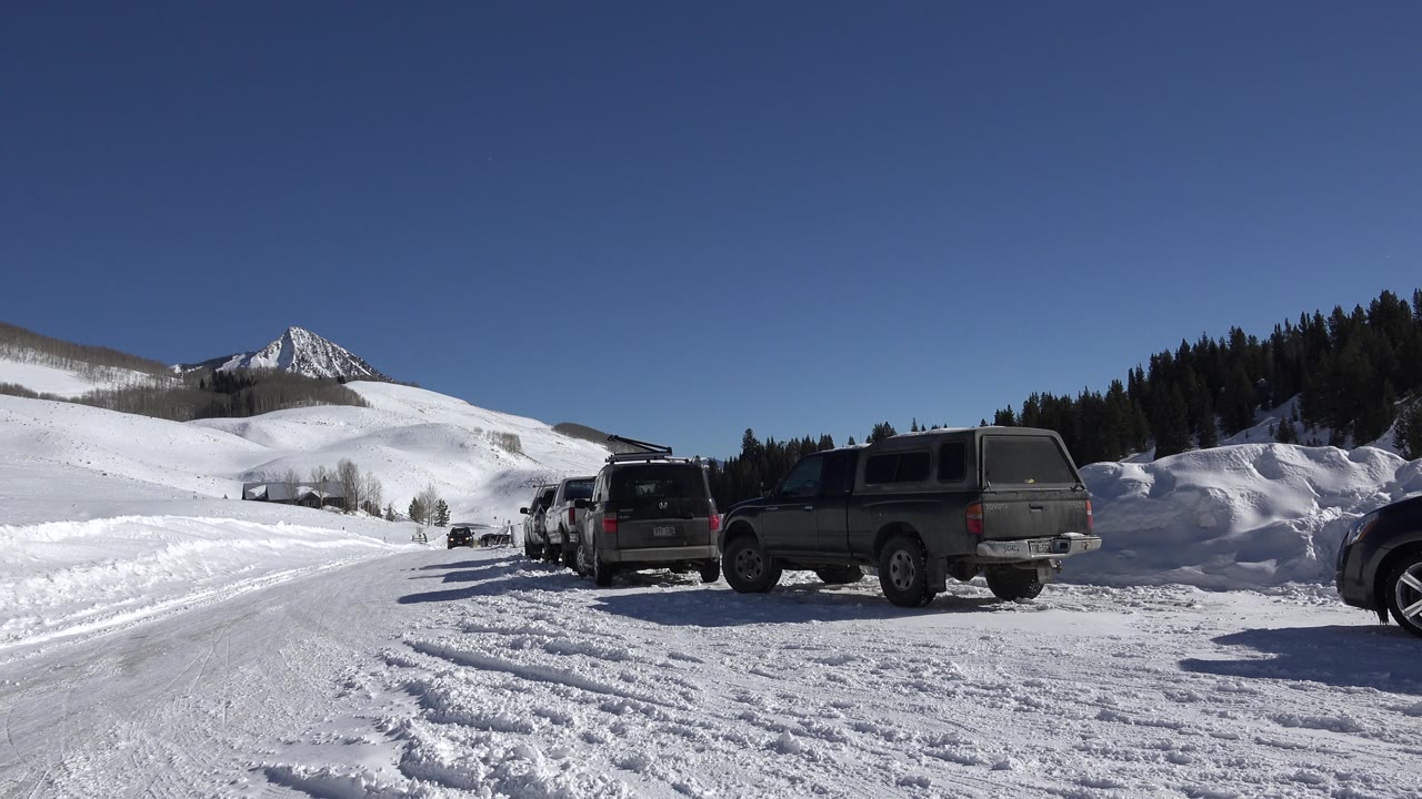 At 0:01, the Crested Butte winter road curves through packed snow toward parked vehicles, rolling white hills, dark trees, a distant pointed mountain, and no visible people.