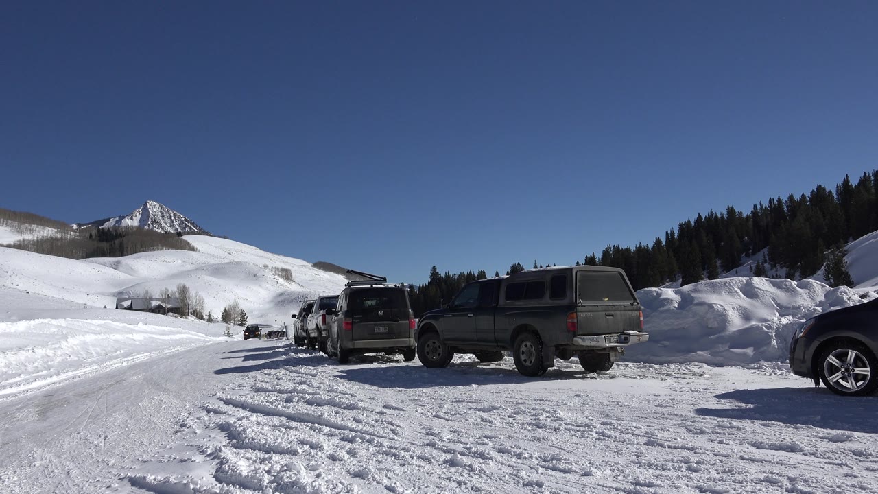 Parked vehicles beside a snow-packed road with bright hills and a mountain in the background.