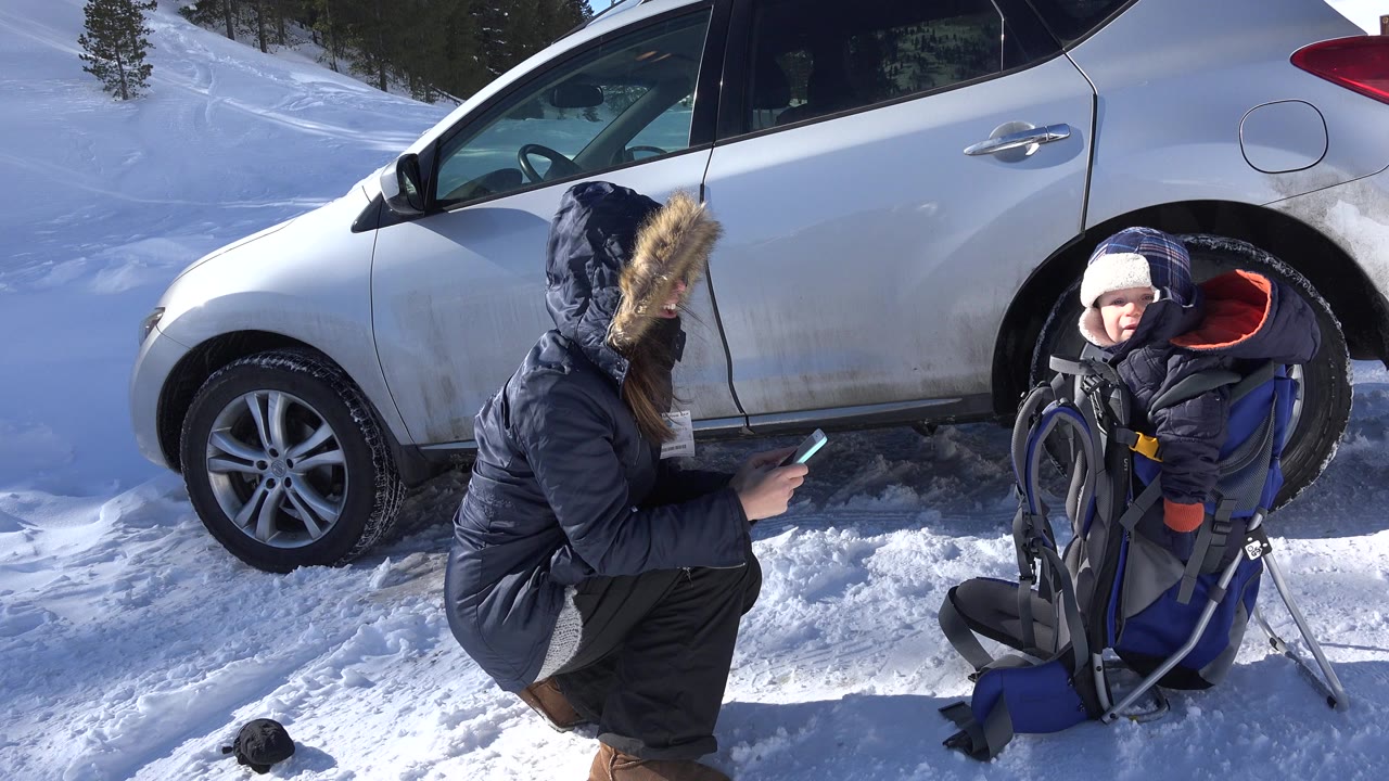 Adult crouching in snow photographing a child in a backpack carrier beside a silver SUV.