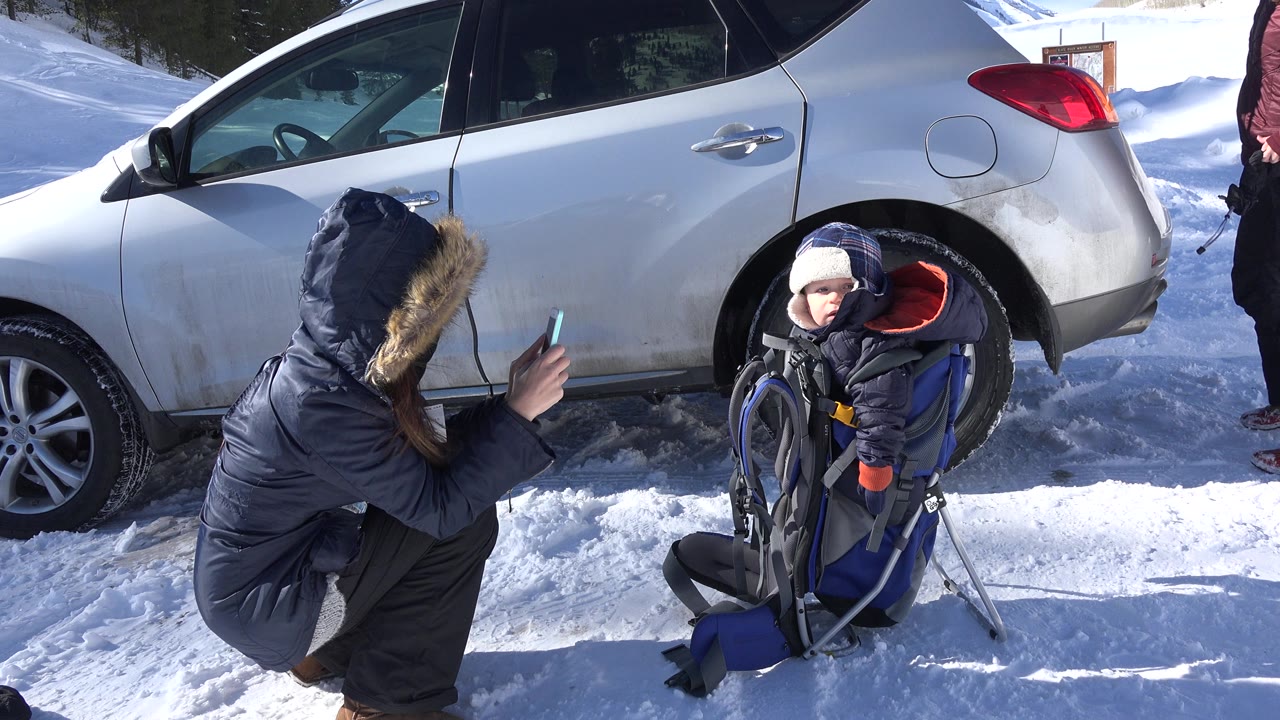 Adult crouching by a child carrier beside a silver SUV in snow.