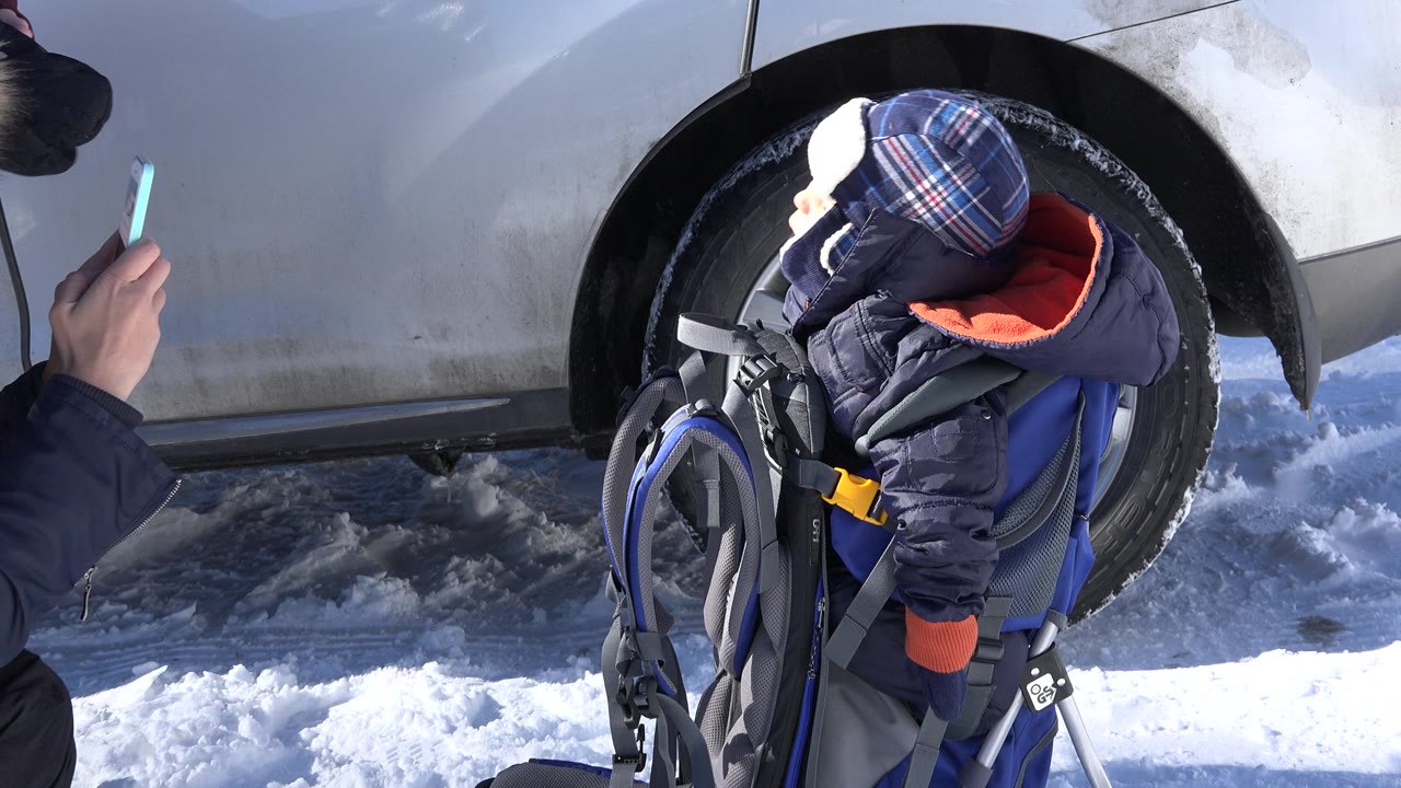 At 0:00 in Crested Butte, Colorado, a bundled baby boy sits in a blue carrier beside a white car wheel on packed snow while an adult hand with a phone is partly visible at the edge.