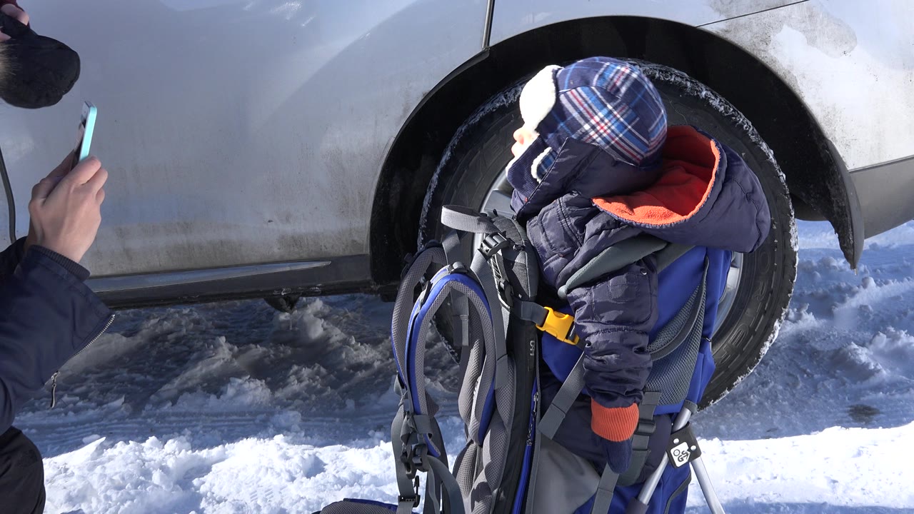 At 0:01 in Crested Butte, Colorado, the baby remains in the carrier near the white car, with snowy ground below and an adult's phone hand near the left edge.