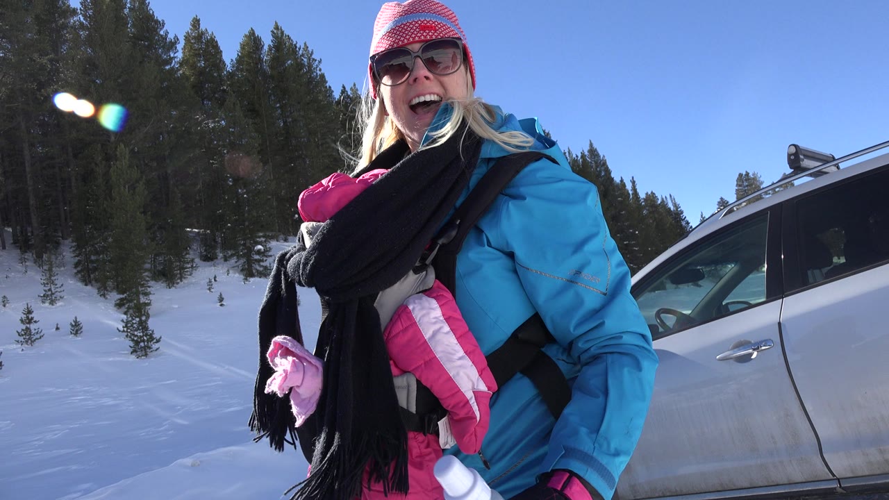 Woman in winter clothes holding a baby carrier beside a silver vehicle in snow.