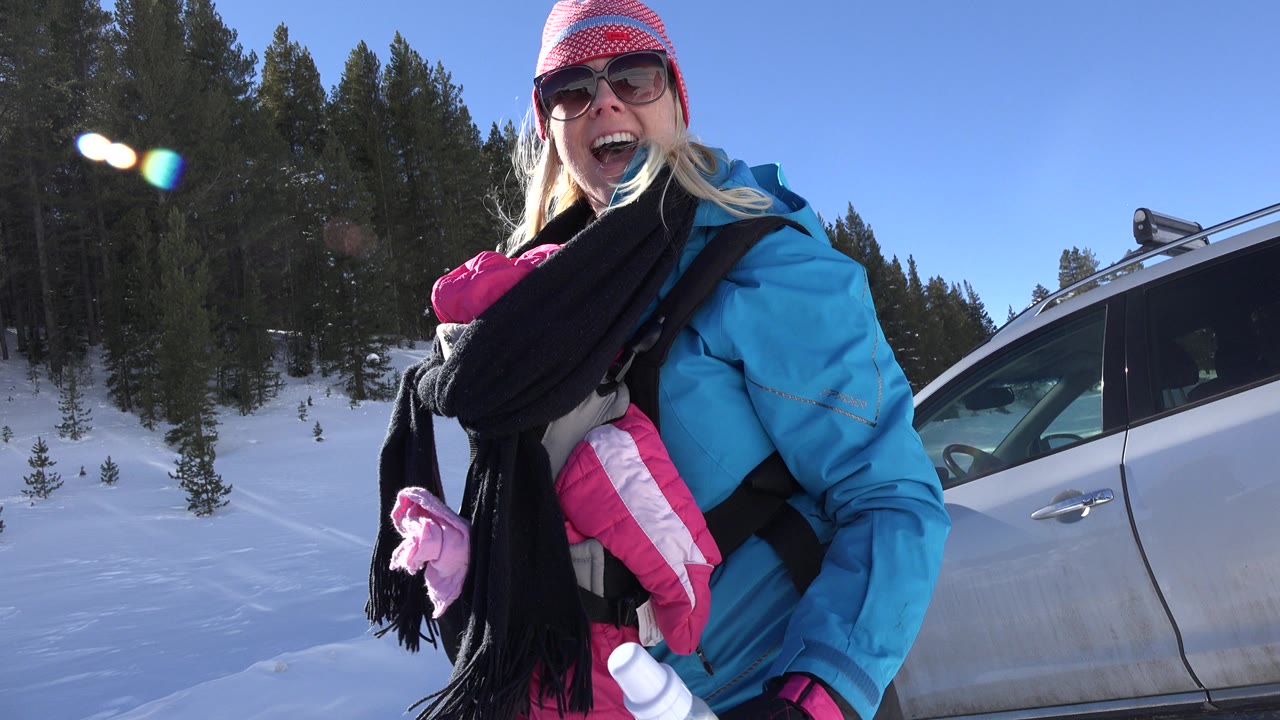 The mother smiles toward the camera while holding the bundled baby in a front carrier, with a silver vehicle and snowy pine slope behind her in Crested Butte, Colorado.