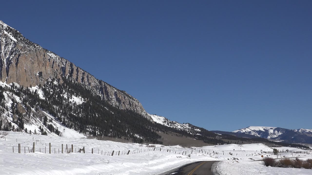 A snow-covered road leads past fence posts toward a rocky mountain slope in Crested Butte, Colorado, with distant ridges and no visible people.