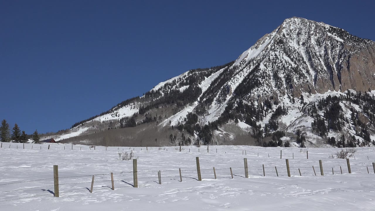 Opening frame of a snow-covered field and scattered fence posts below a large snow-streaked mountain in Crested Butte, Colorado, with no visible people.