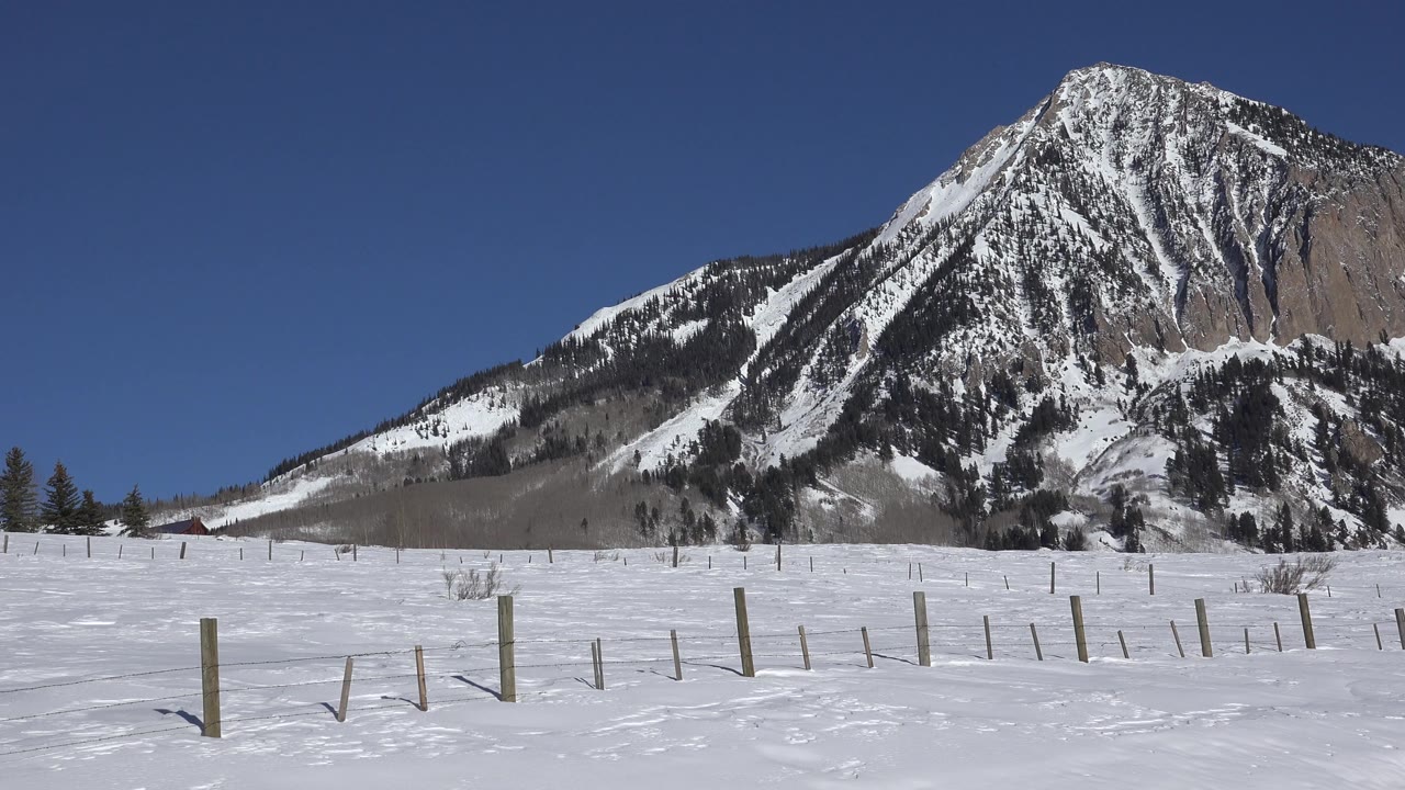 Wide view of the Crested Butte, Colorado snowfield with a low fence line in front of a rocky mountain face and clear blue sky, with no visible people.
