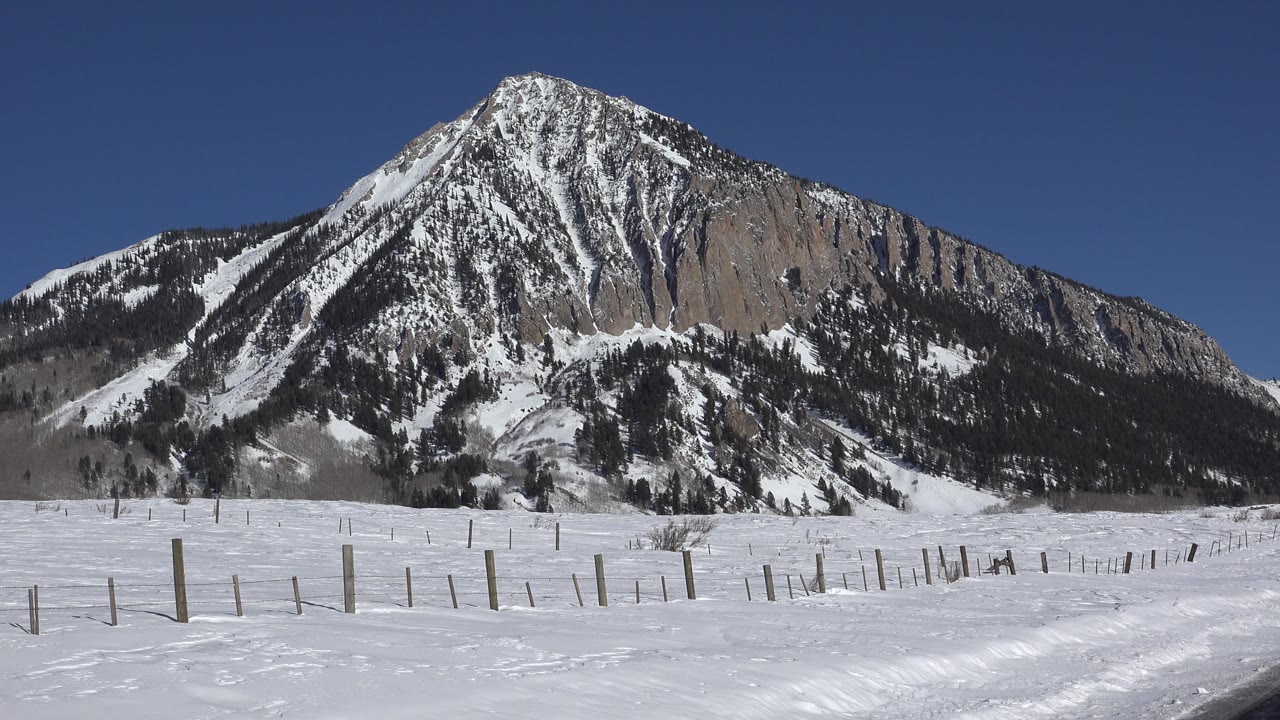 Wide winter view in Crested Butte, Colorado with fence posts across a snow-covered foreground, a steep mountain in back, and no visible people.