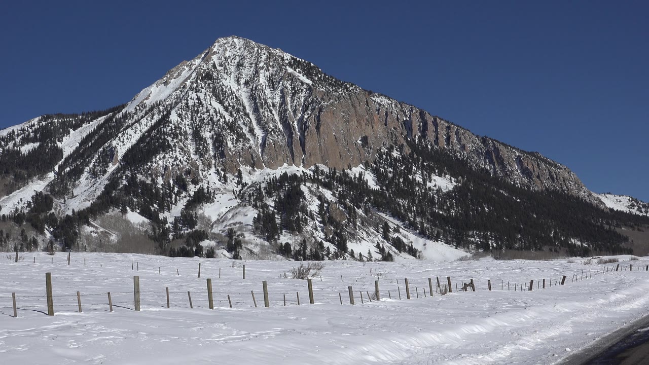 Snowy open land and mountain slope with a road edge beginning at right.