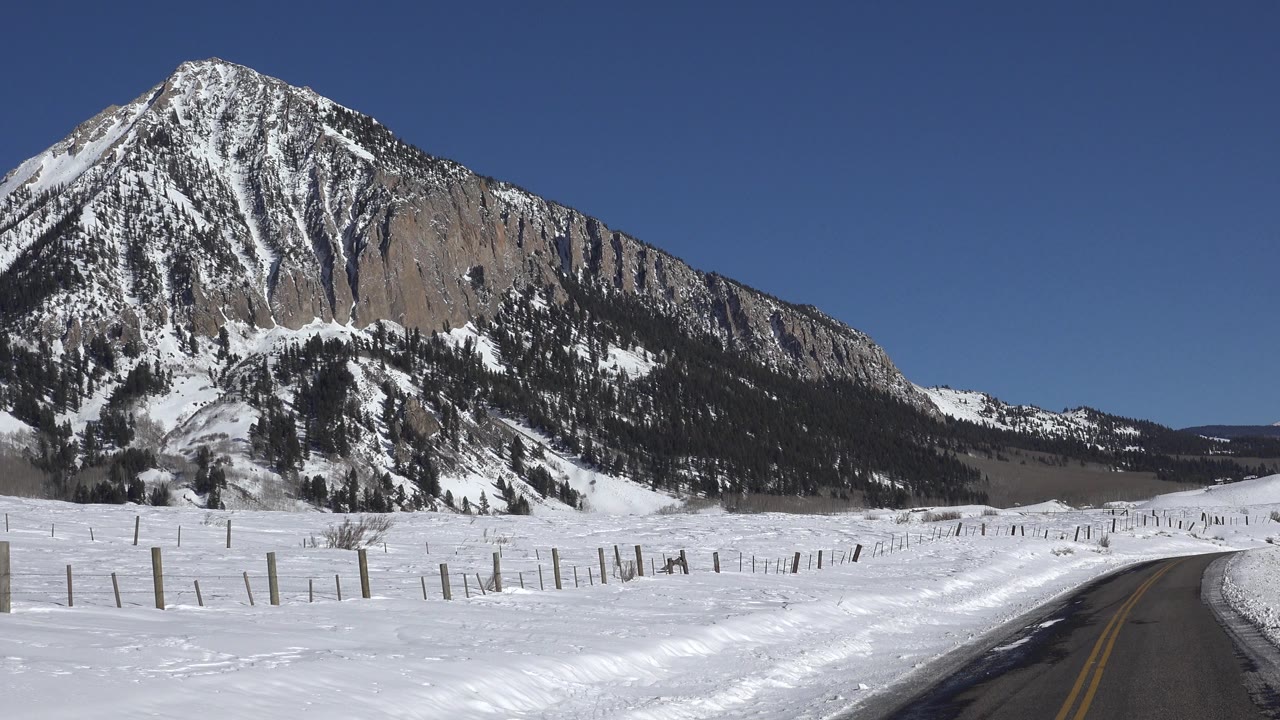 Opening frame of a snowy two-lane road in Crested Butte, Colorado, with fence posts across the snowfield, a steep mountain face at left, and no visible people.