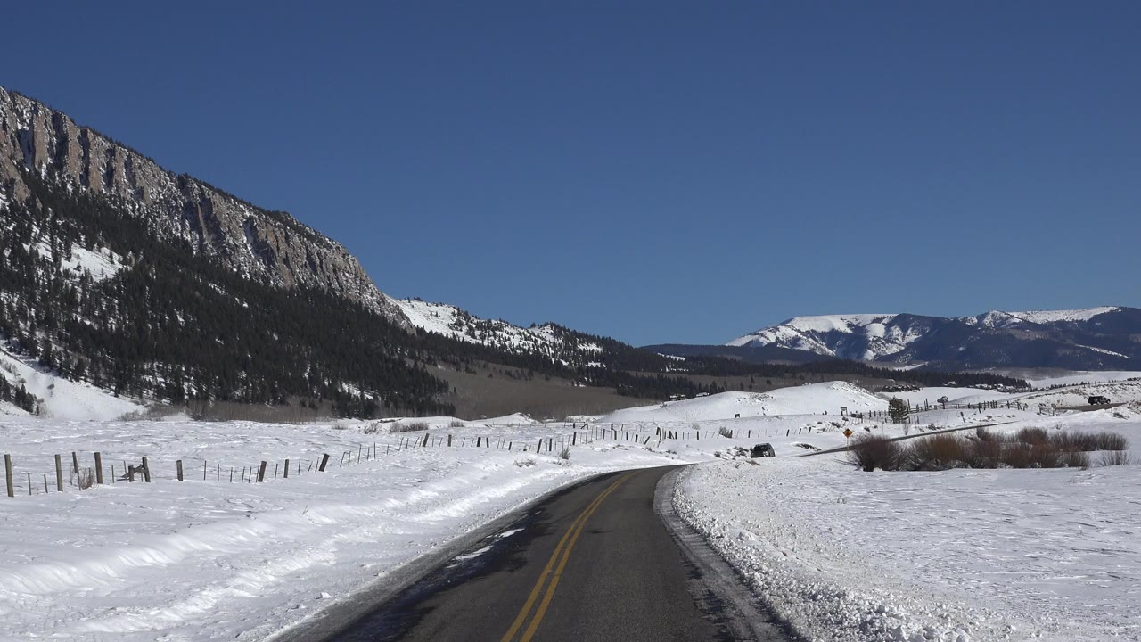 Opening frame of a Crested Butte, Colorado winter road curving through snow fields beneath a clear blue sky, with no visible people.