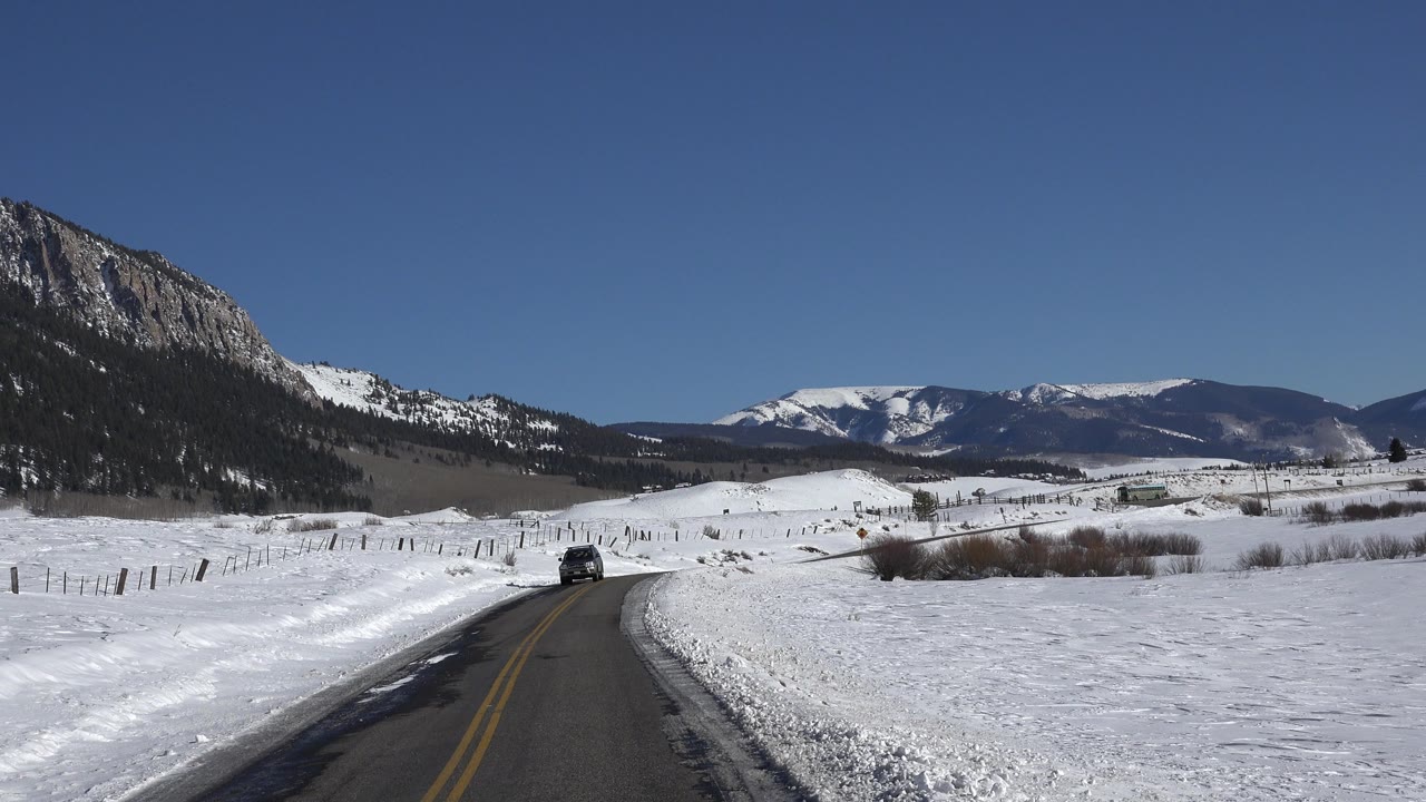 Opening frame of a Crested Butte winter road bordered by snow fields, distant mountains, and a clear blue sky, with no visible people.