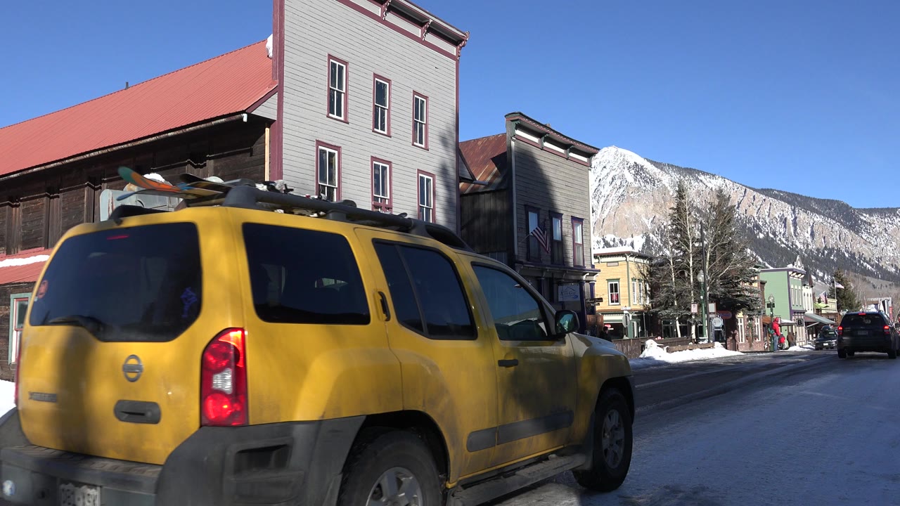 A yellow SUV fills the left foreground on a snowy Crested Butte street, with storefront buildings, a distant mountain, blue sky, and no visible people.