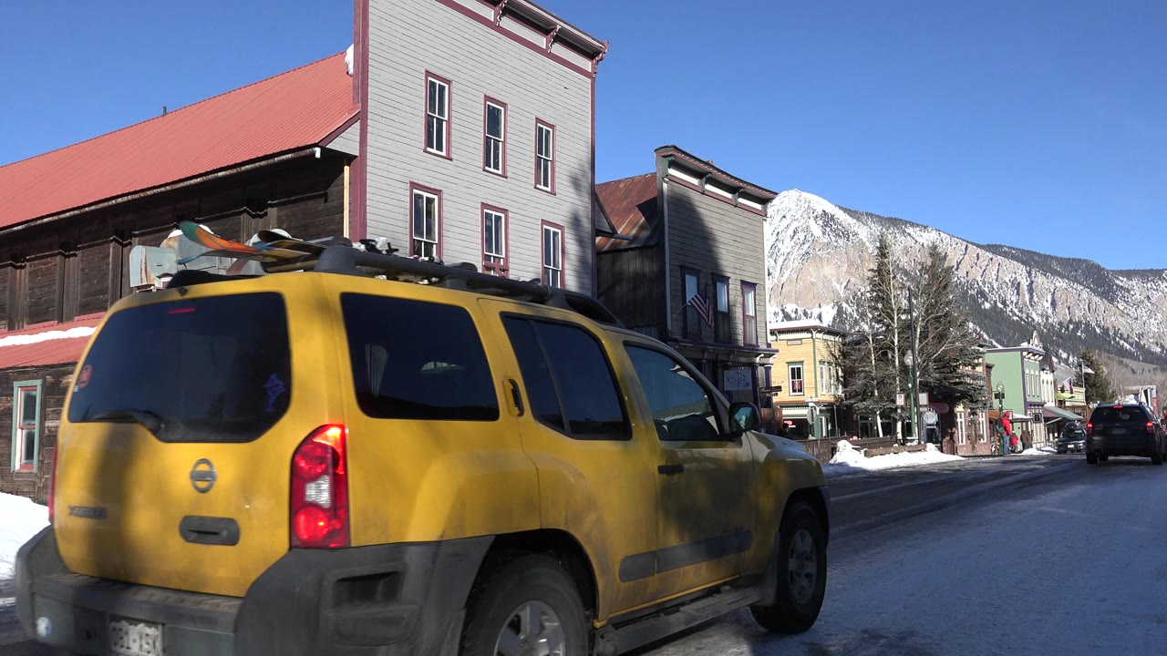 The yellow SUV remains large in the foreground as it continues along the snow-lined Crested Butte street, with red-roofed buildings and the mountain still visible, no visible people.