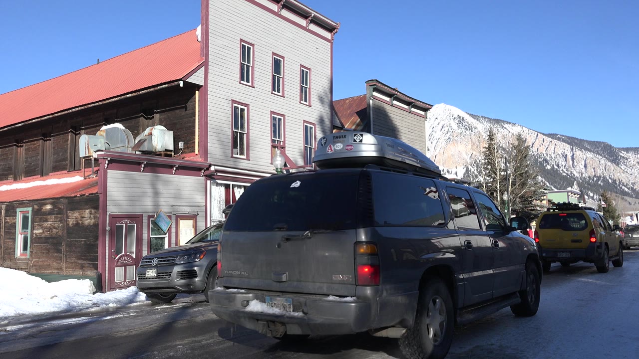 A gray SUV fills the foreground on a Crested Butte street, with parked cars, snowbanks, older buildings, blue sky, and a snow-covered mountain behind it, with no visible people.