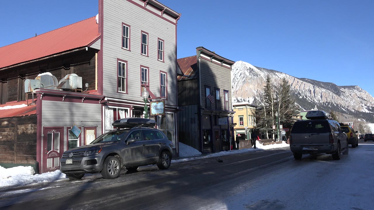 Snowy town street with parked SUV, storefronts, and mountains.