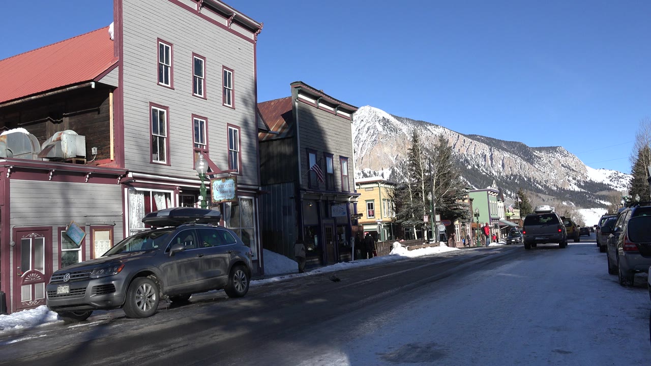 Snow-lined street with parked cars, storefront buildings, and mountains in the background.