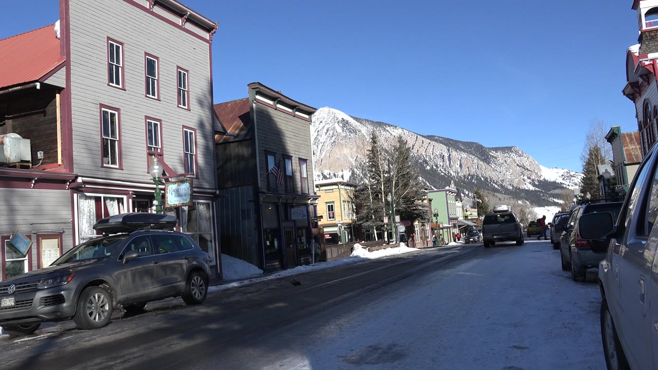 Snowy town street with parked cars and mountains in the distance.