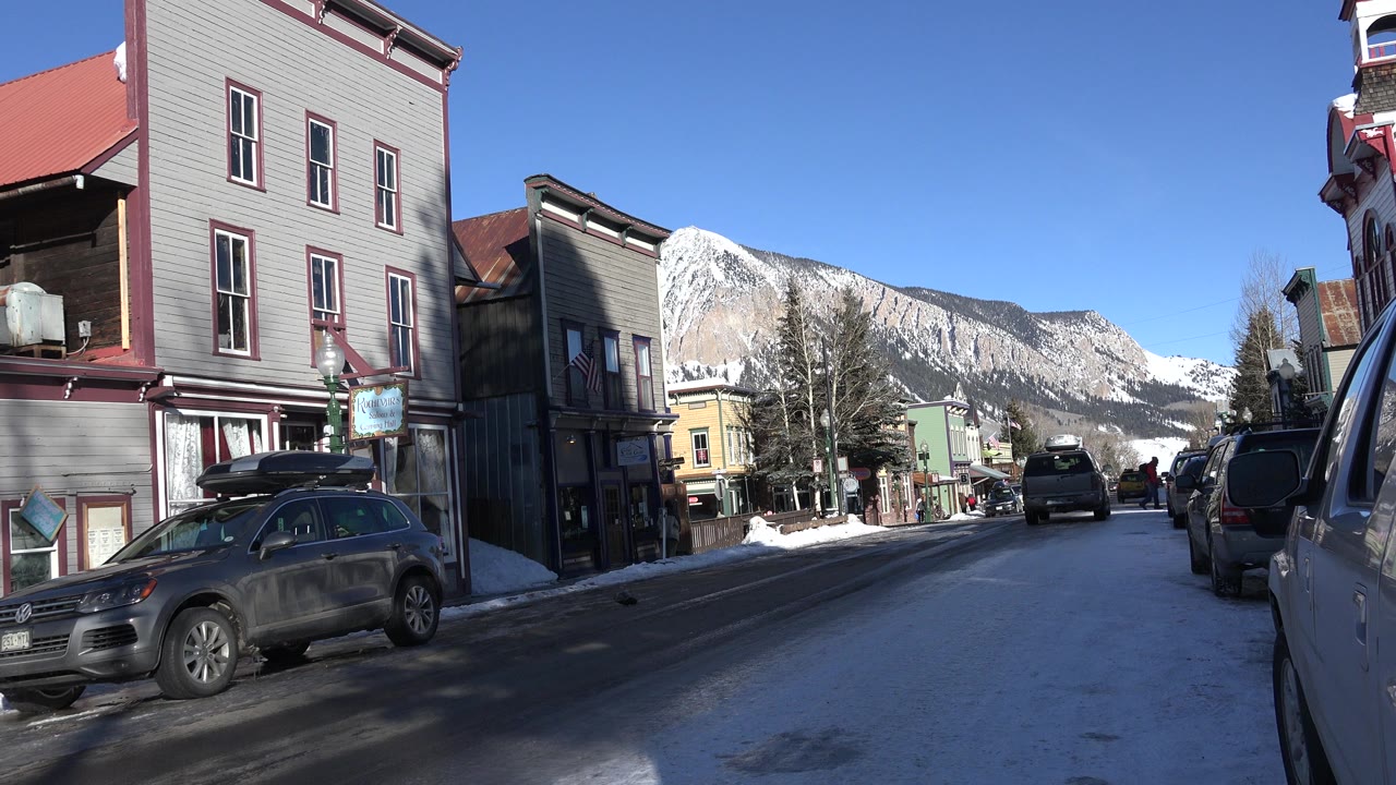 Crested Butte street view one second in, with a dark parked car on the left, a vehicle edge close on the right, old storefront-style buildings, snow patches, mountains, and no visible people.