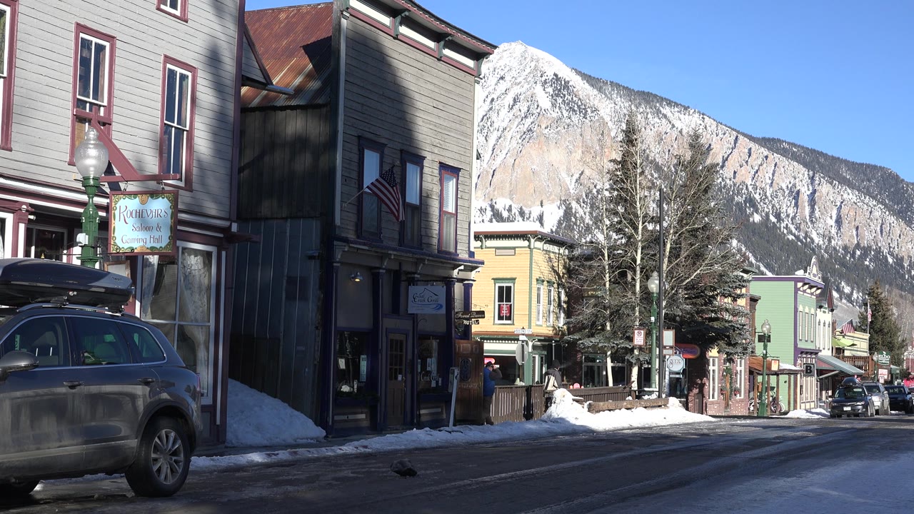 Crested Butte storefronts and a parked SUV frame the lower street while a sunlit snow-covered mountain ridge rises behind the town, with no clearly visible people.