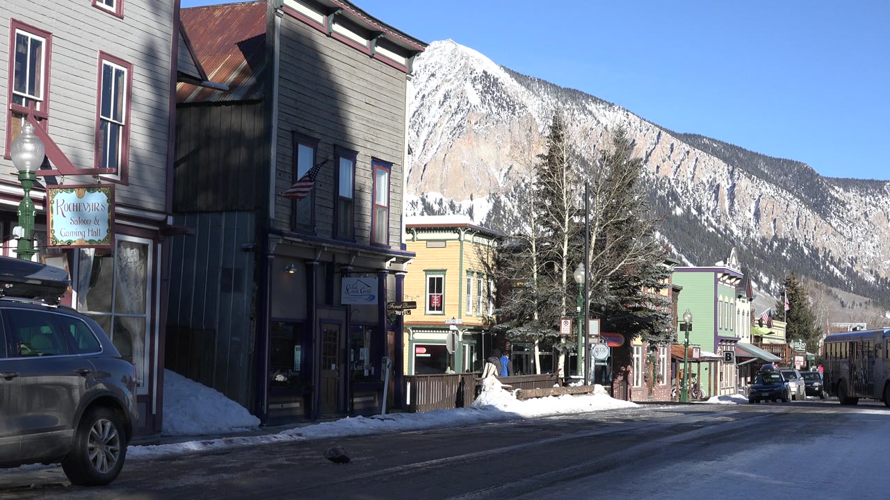 Crested Butte, Colorado street view at the start of the clip, with shaded storefronts at left, a vehicle at the curb, snowbanks, blue sky, and snow-dusted mountains behind, with no visible people.