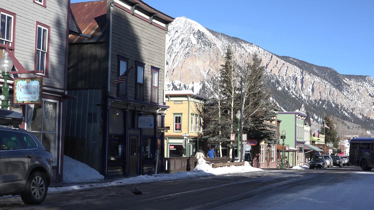 Mountain town road with storefronts, parked vehicles, snow, and a steep ridge.