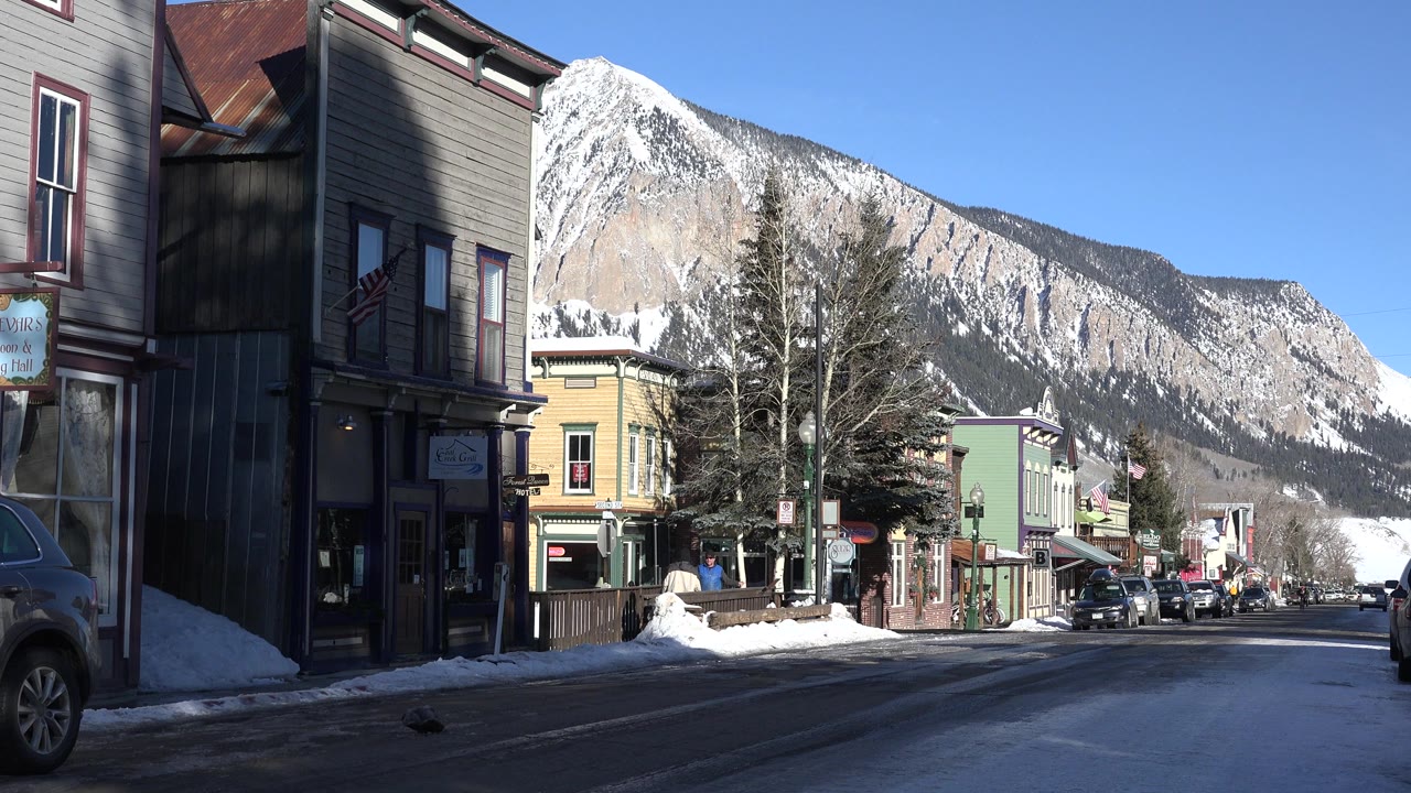 Opening frame of a Crested Butte street with snowbanks, parked vehicles, storefront buildings, bare trees, a mountain backdrop, and no visible people.