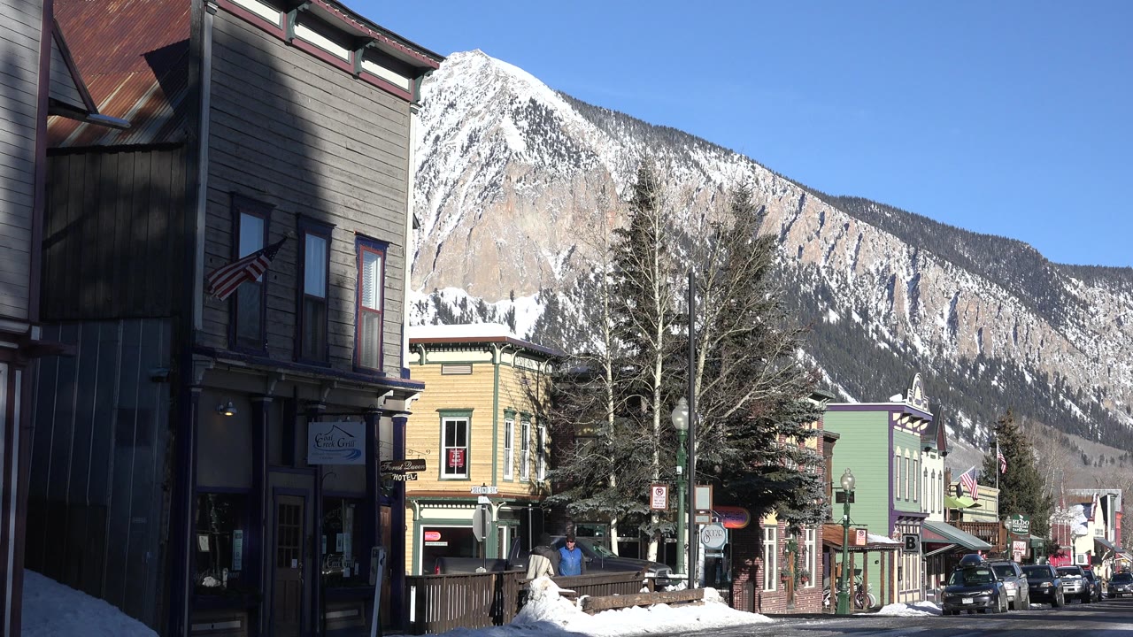 Crested Butte street view at the start of the clip, with shaded buildings on the left, snowbanks, parked cars, bare trees, American flags, and a snow-covered mountain behind town.