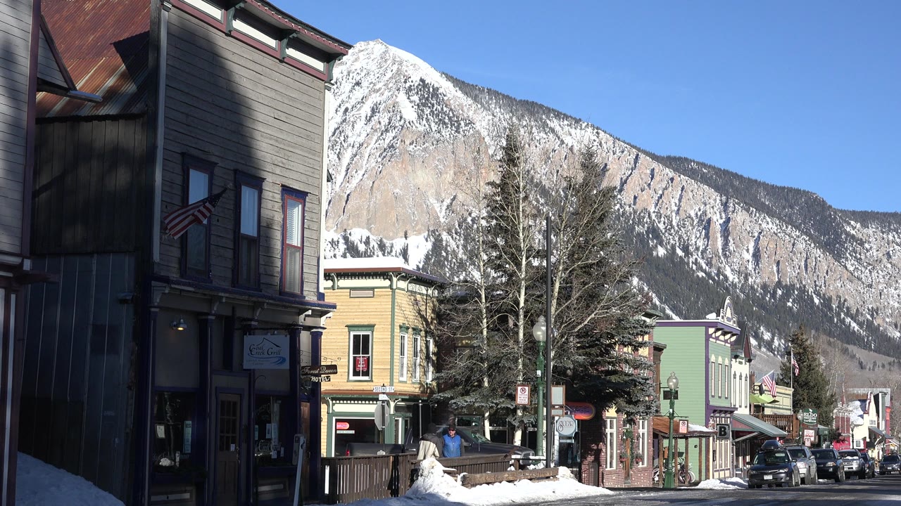 Crested Butte winter street frame with the dark building edge at left, painted storefronts near the center, several cars along the road, small pedestrians, and a steep snowy mountain in sunlight.