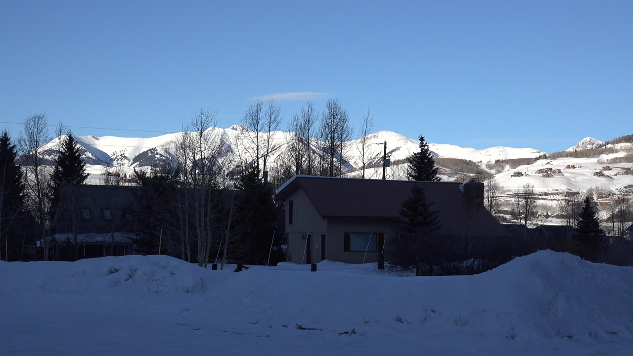 Snowy yard with a house, trees, and mountains under a blue sky.