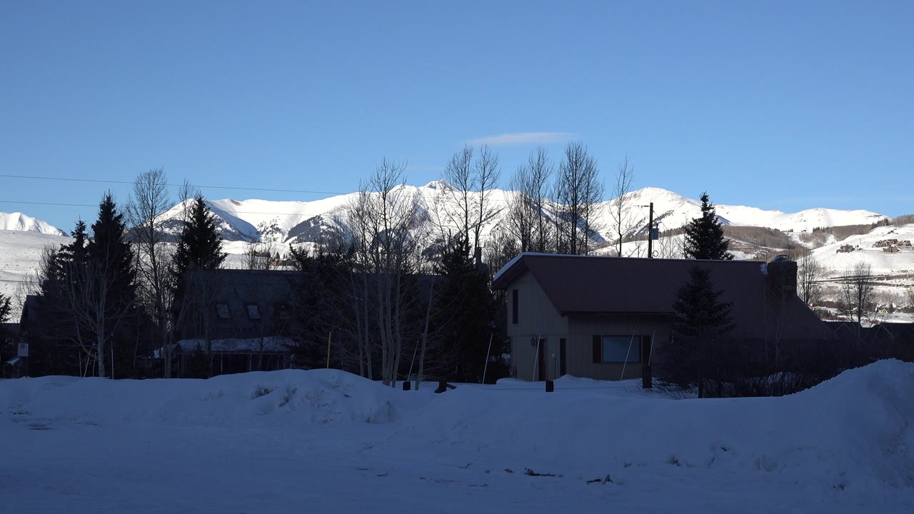 Snow-covered Crested Butte mountains stretch across the background while shaded homes, bare trees, evergreens, and snowbanks fill the lower frame, with no visible people.