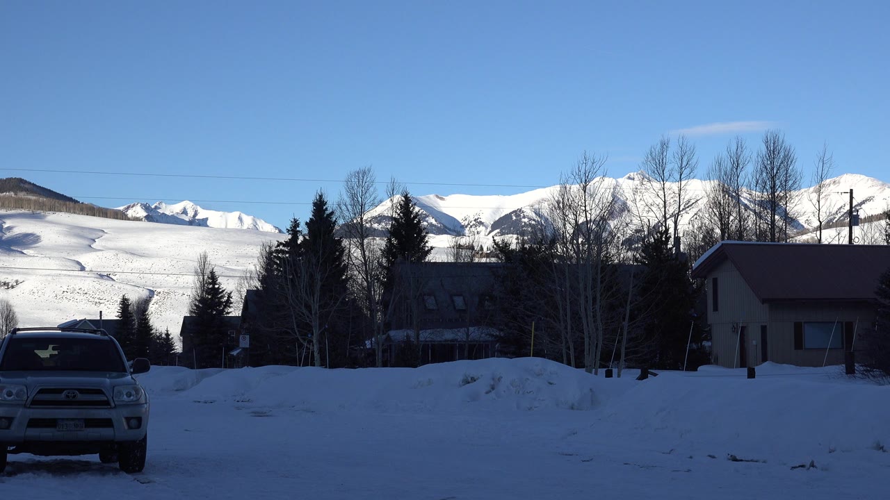 Pickup truck in snow with houses, trees, and snowy mountains behind it.